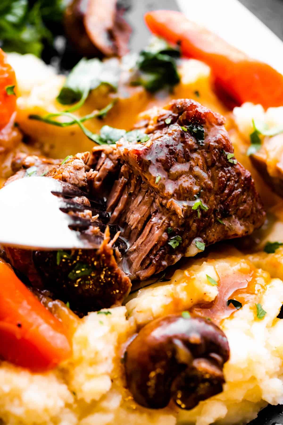 A fork cutting into a pot roast set atop mashed potatoes and surrounded with carrots.