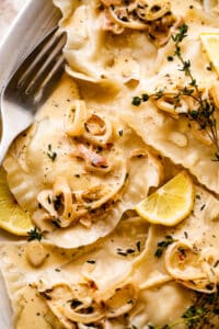 close up shot of lobster ravioli arranged on a plate and garnished with cream sauce, lemon slices, and thyme.