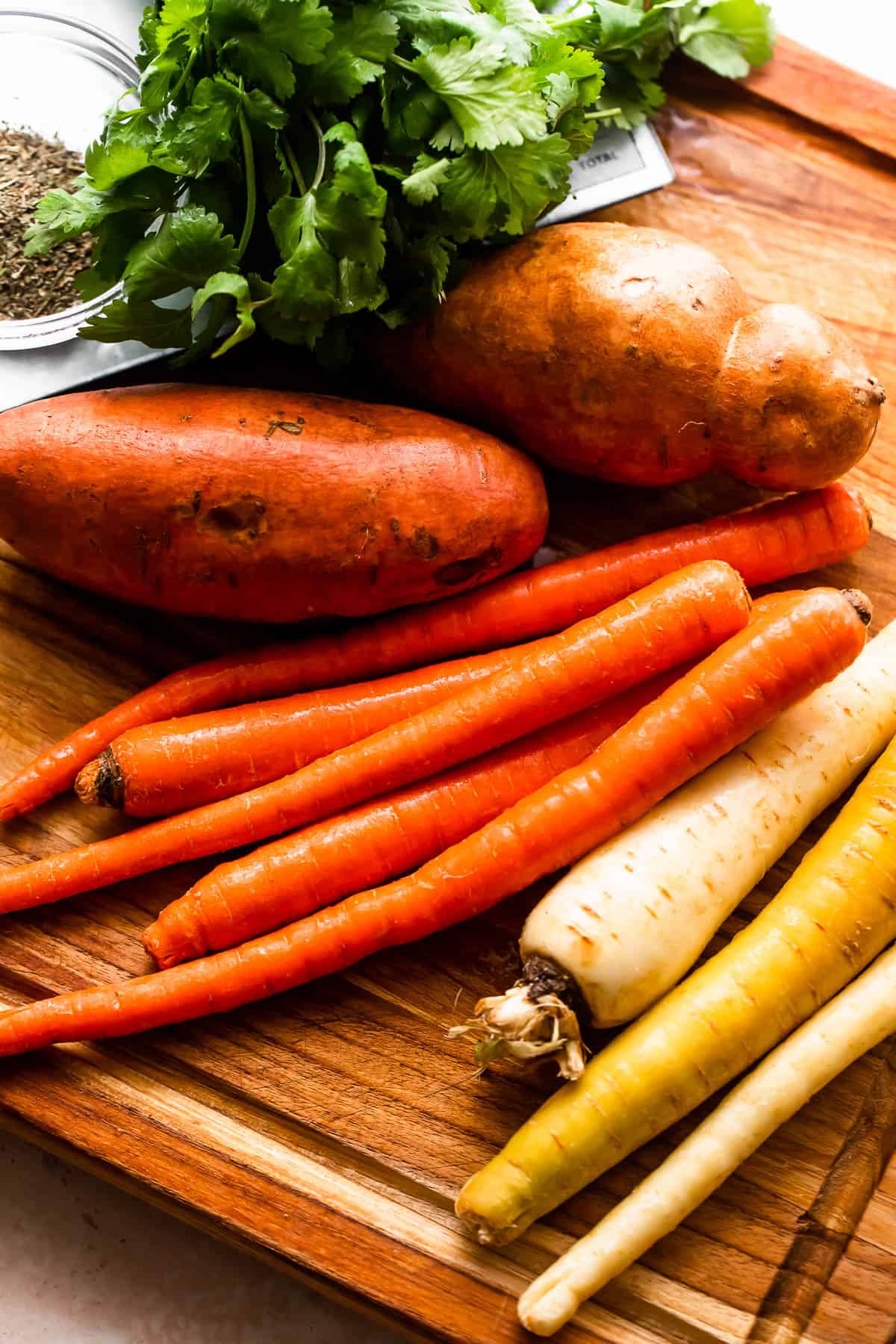 whole sweet potatoes, carrots, parsnips. and parsley on a wooden chopping board
