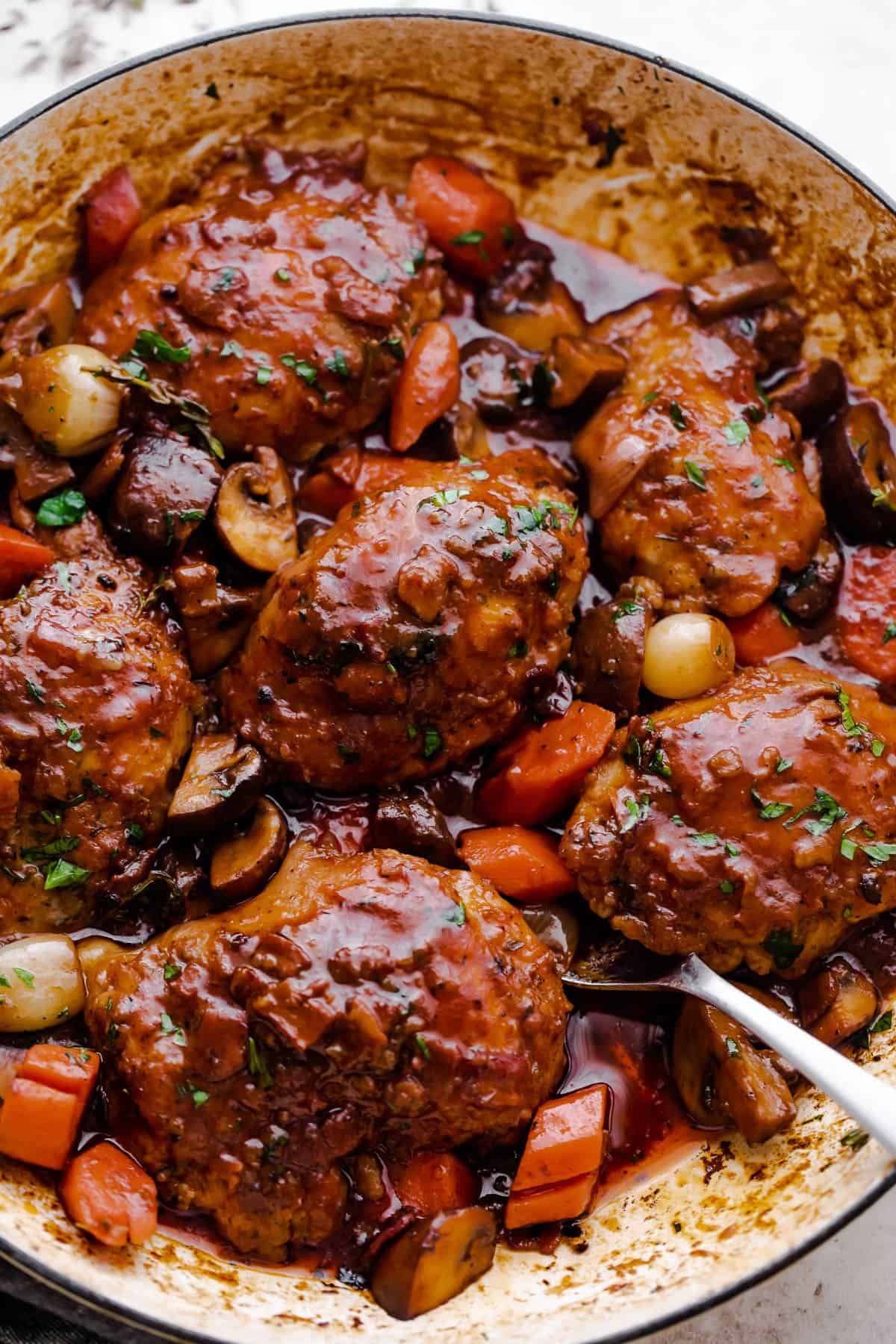 overhead shot of Coq au Vin style cooked chicken thighs in a braiser with chopped carrots, pearl onions, and mushrooms.