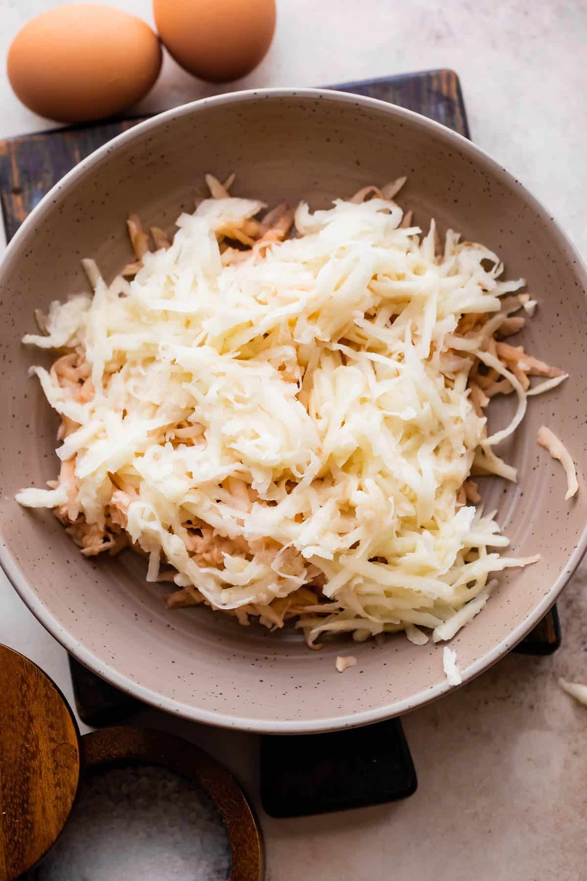 grated potato and onion mixture in a brown bowl with two brown eggs placed next to the bowl.