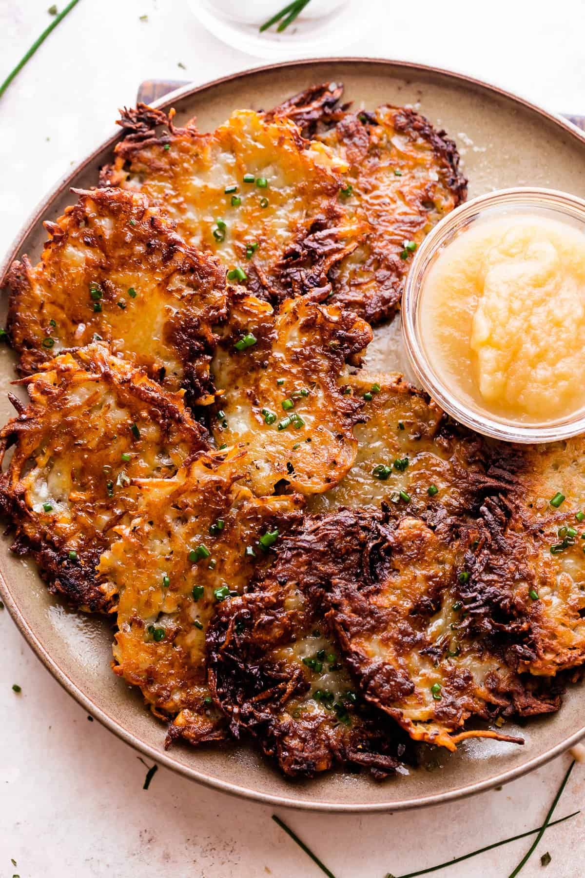 potato latkes arranged on a light brown plate with a small bowl of applesauce placed next to the latkes, and a bowl of sour cream near the plate.