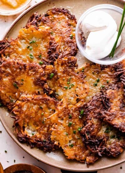 potato latkes arranged on a light brown plate with a small bowl of sour cream placed next to the latkes, and a bowl of applesauce near the plate.