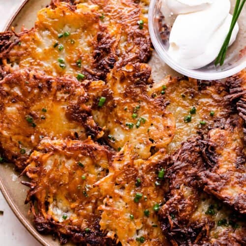 potato latkes arranged on a light brown plate with a small bowl of sour cream placed next to the latkes, and a bowl of applesauce near the plate.