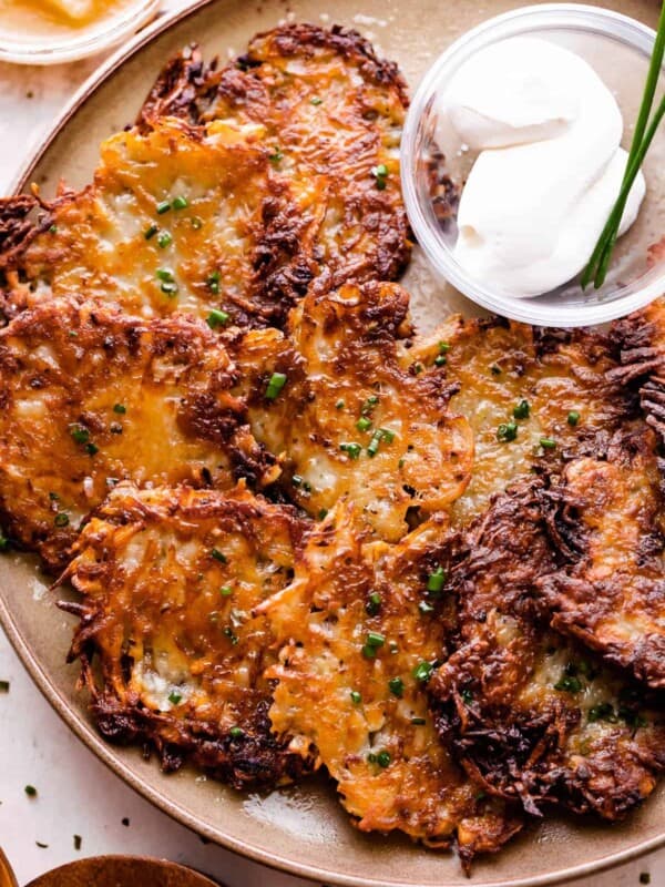 potato latkes arranged on a light brown plate with a small bowl of sour cream placed next to the latkes, and a bowl of applesauce near the plate.