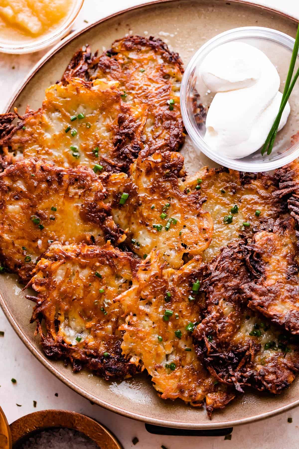 potato latkes arranged on a light brown plate with a small bowl of sour cream placed next to the latkes, and a bowl of applesauce near the plate.