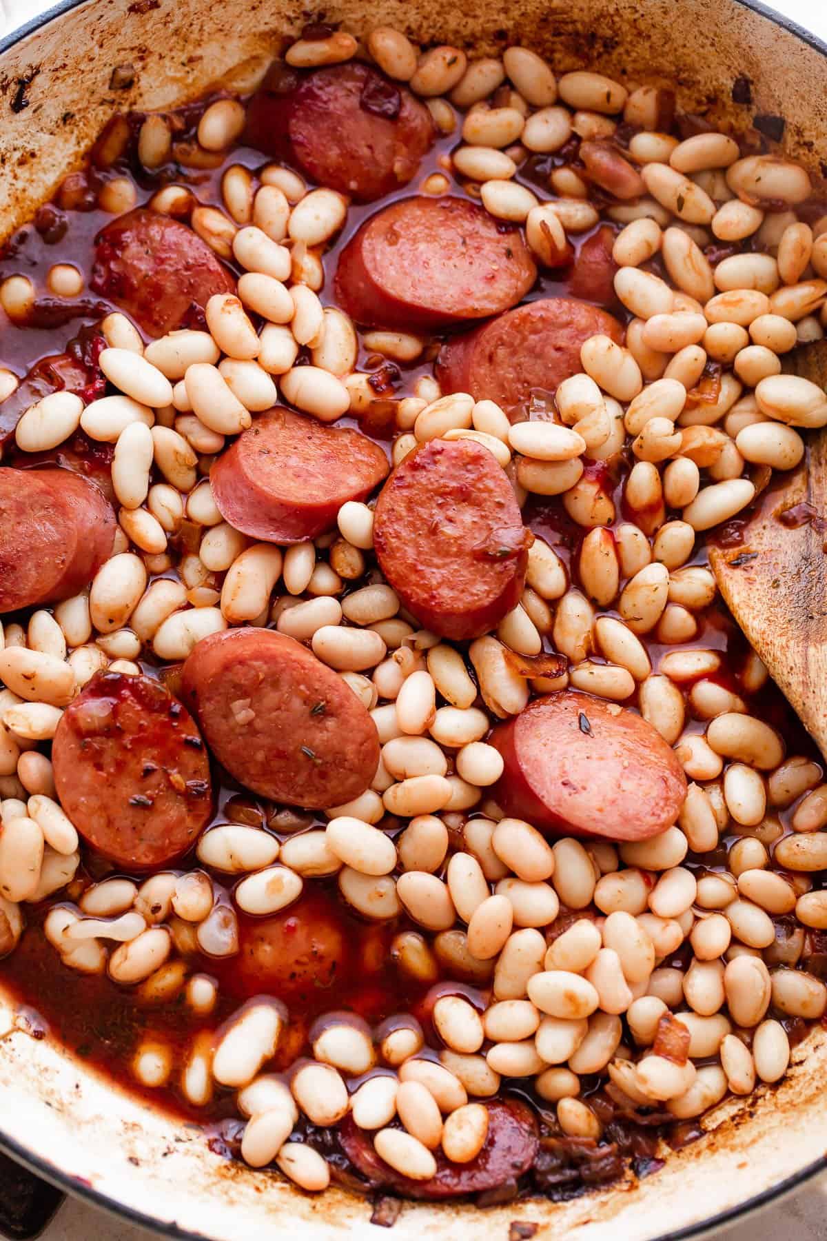 Smoked sausage slices and white beans cooking in a white skillet.