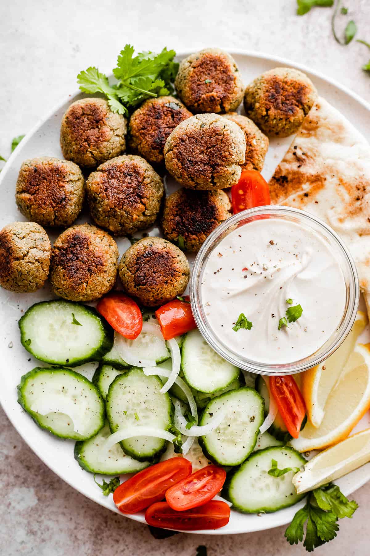 overhead shot of baked falafel, cucumber slices, tomatoes, lemons, and tahini sauce all arranged on a large round plate