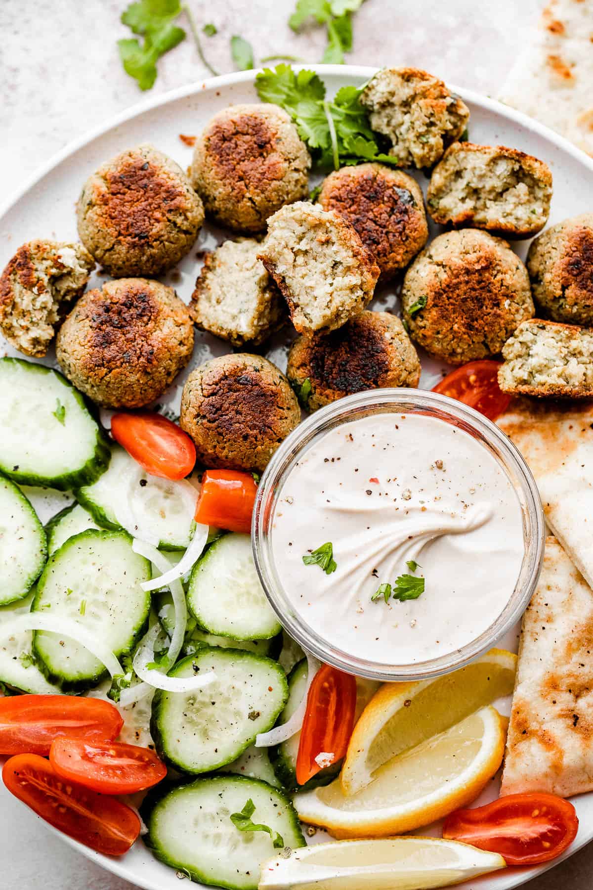 overhead shot of baked falafel, cucumber slices, tomatoes, lemons, and tahini sauce all arranged on a large round plate