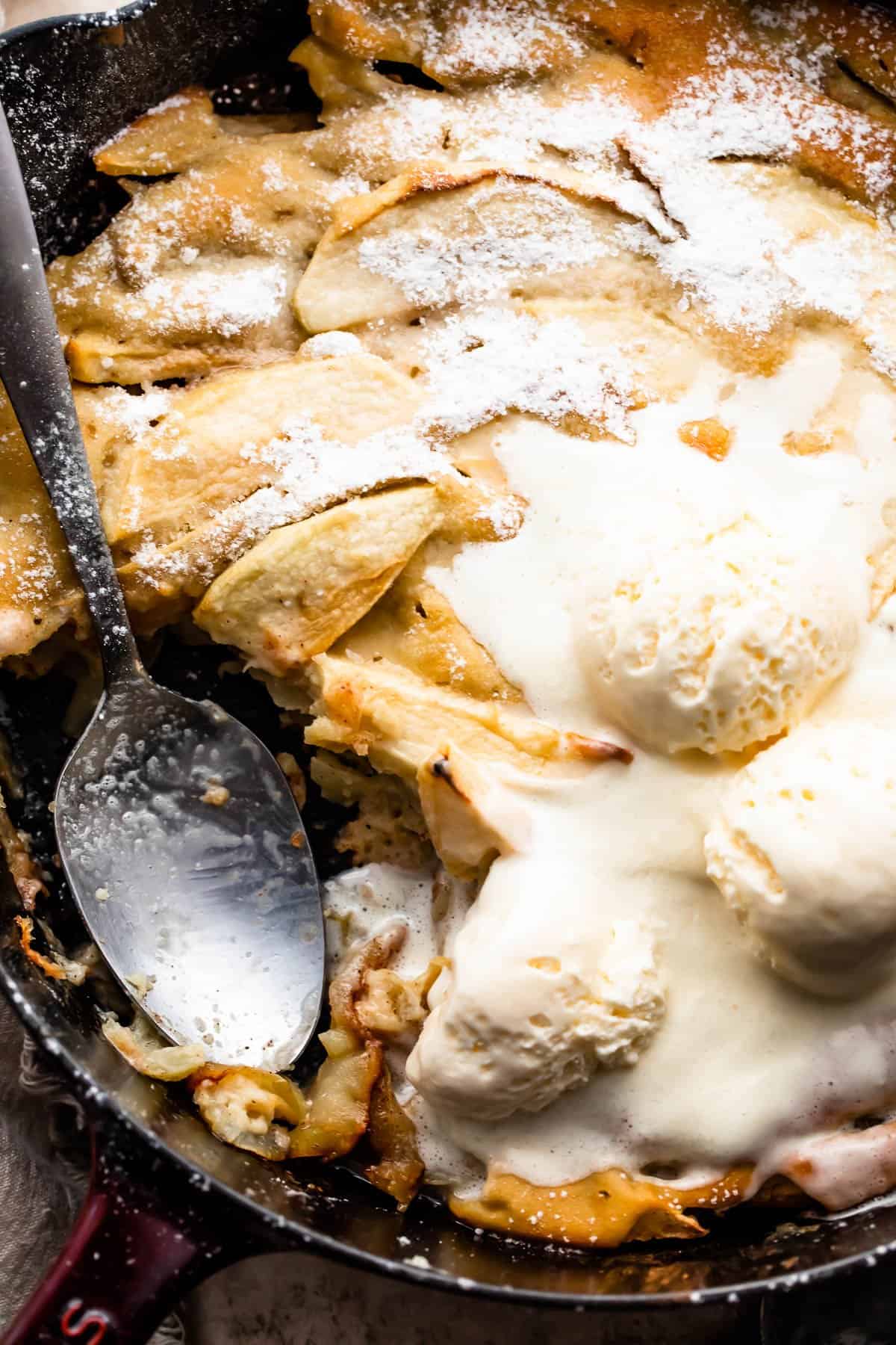 Close-up overhead shot of a healthy apple cobbler in a skillet, topped with three scoops of vanilla ice cream, and spooning out the cobbler with a big spoon.