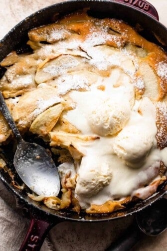 overhead shot of healthy apple cobbler in a skillet, topped with three scoops of vanilla ice cream, and spooning out the cobbler with a big spoon.