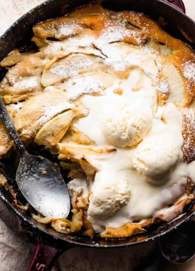 overhead shot of healthy apple cobbler in a skillet, topped with three scoops of vanilla ice cream, and spooning out the cobbler with a big spoon.