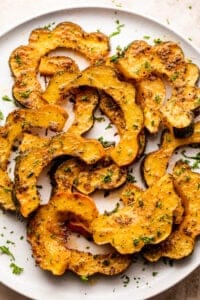 roasted acorn squash cut into half-moon slices, set on a white plate and seasoned with oil and herbs.