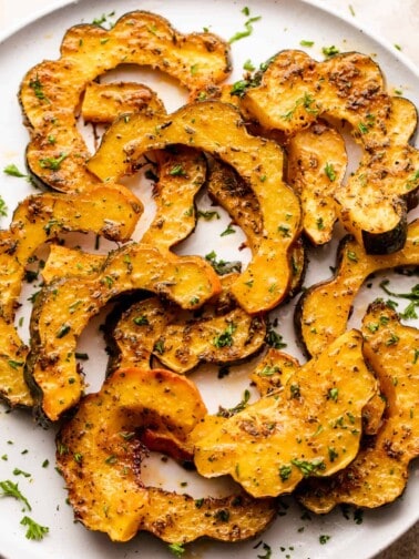 roasted acorn squash cut into half-moon slices, set on a white plate and seasoned with oil and herbs.
