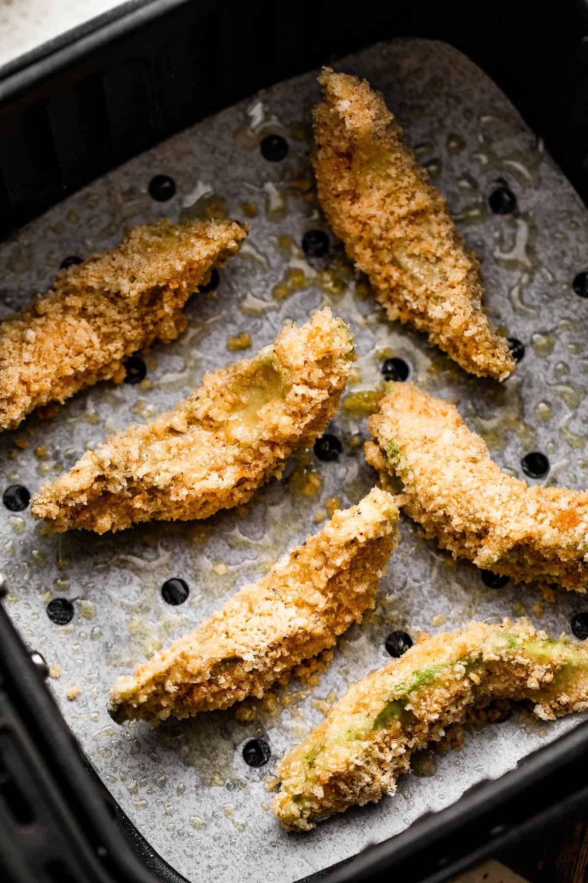 Overhead shot of breaded avocado slices arranged in a black air fryer basket.