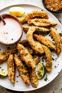 overhead shot of air fried avocado fries arranged on a white plate with a small bowl of ranch dipping sauce also served on the plate.