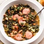 overhead shot of a white bowl filled with black eyed peas, kale, sliced andouille sausage, and broth. Bread slices are placed near the top right of the soup bowl.