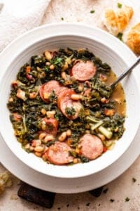 overhead shot of a white bowl filled with black eyed peas, kale, sliced andouille sausage, and broth. Bread slices are placed near the top right of the soup bowl.