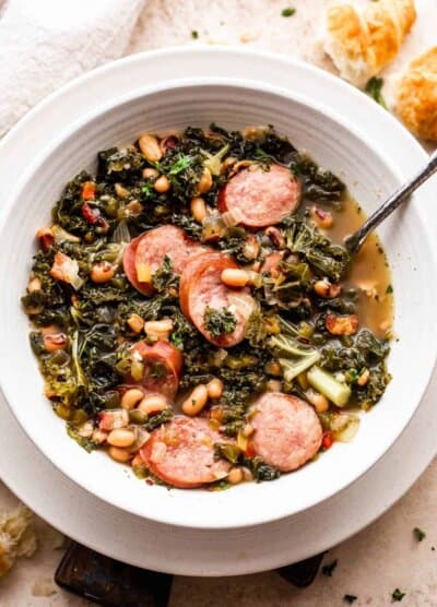 overhead shot of a white bowl filled with black eyed peas, kale, sliced andouille sausage, and broth. Bread slices are placed near the top right of the soup bowl.