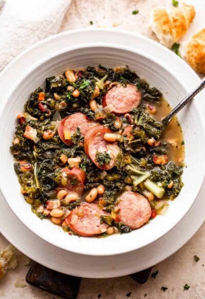 overhead shot of a white bowl filled with black eyed peas, kale, sliced andouille sausage, and broth. Bread slices are placed near the top right of the soup bowl.