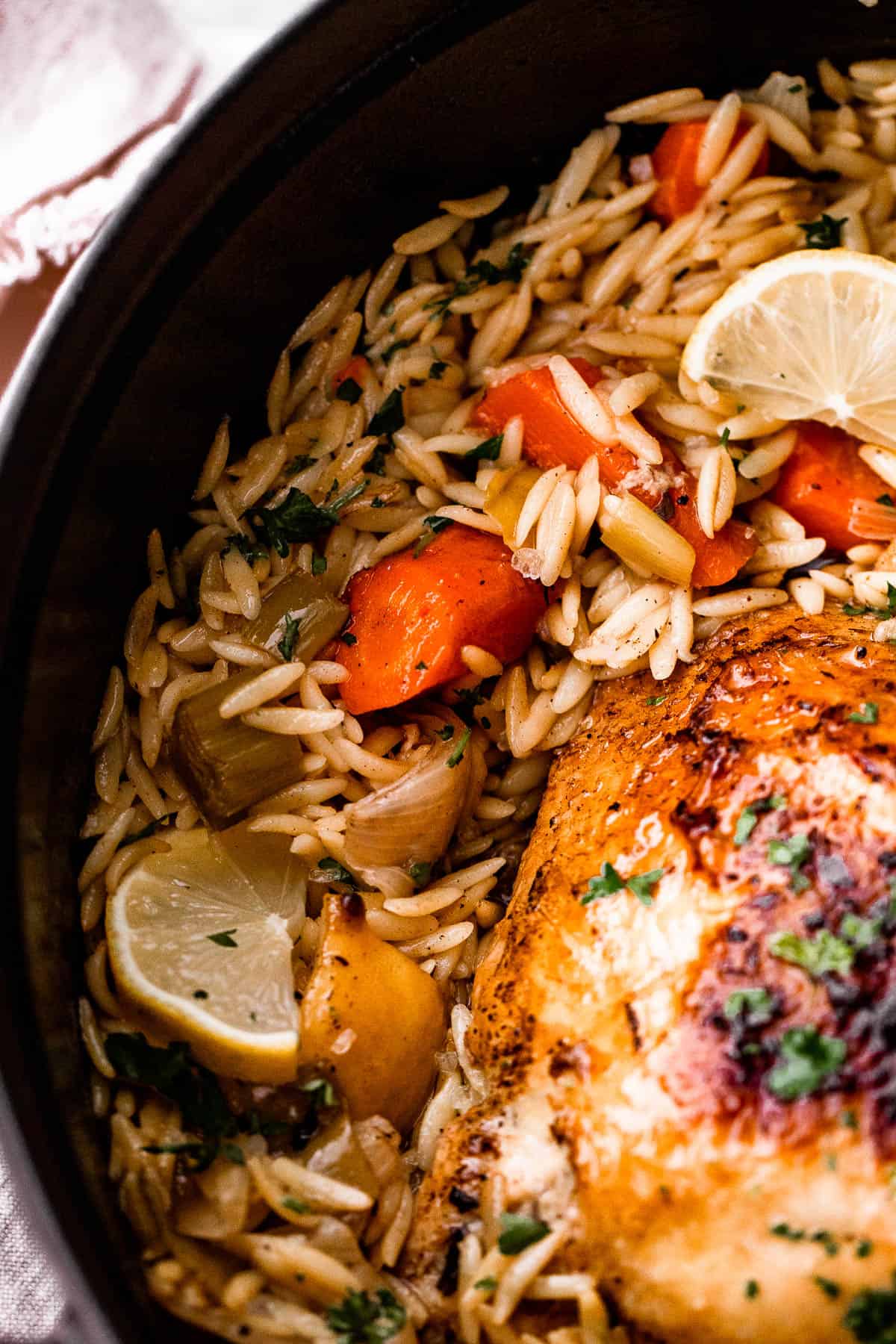 close up shot of cooked orzo mixed in with carrots and lemon wedges, plus a whole roast chicken set on top of the orzo.