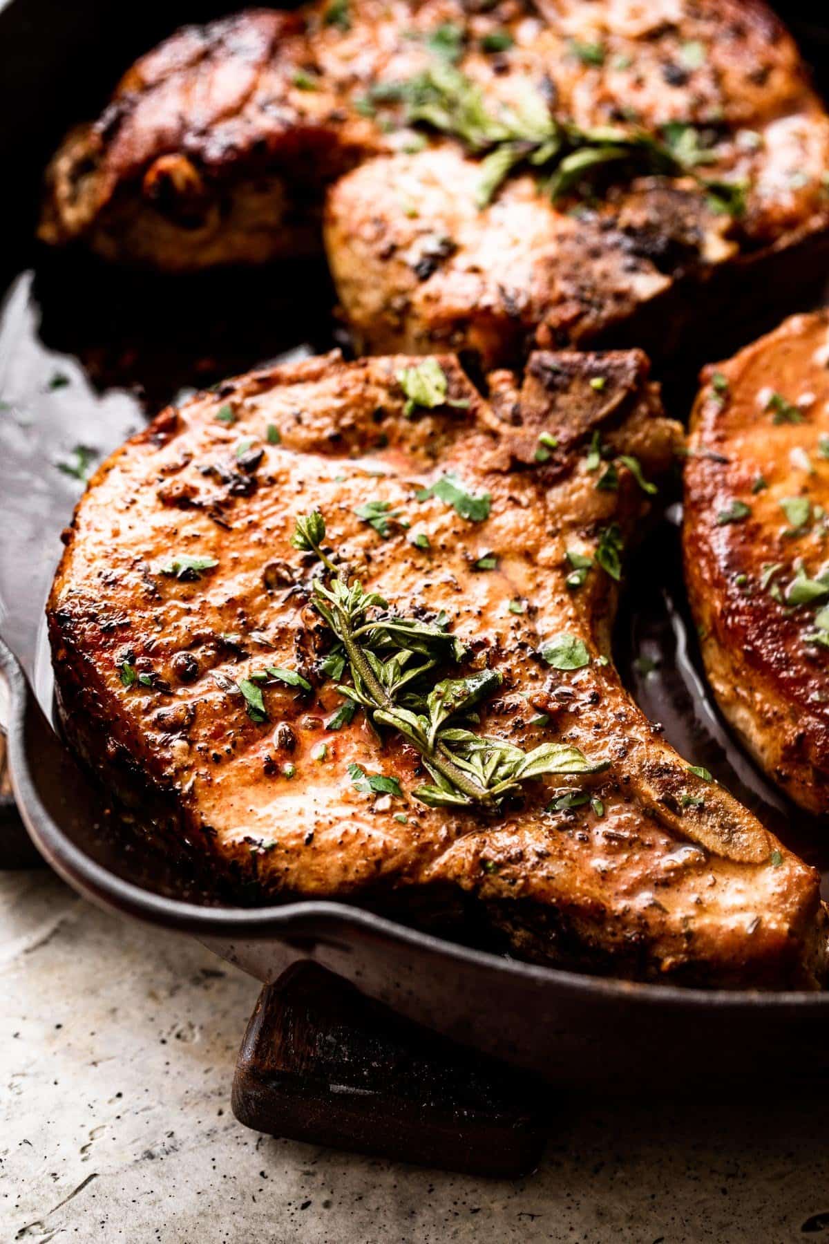 side shot of browned pork chops arranged in a skillet and garnished with fresh herbs