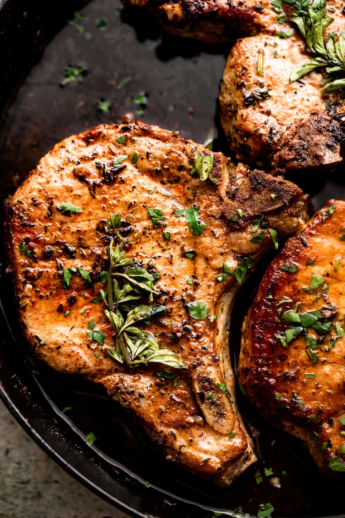 overhead close up shot of browned pork chops arranged in a skillet and garnished with fresh herbs