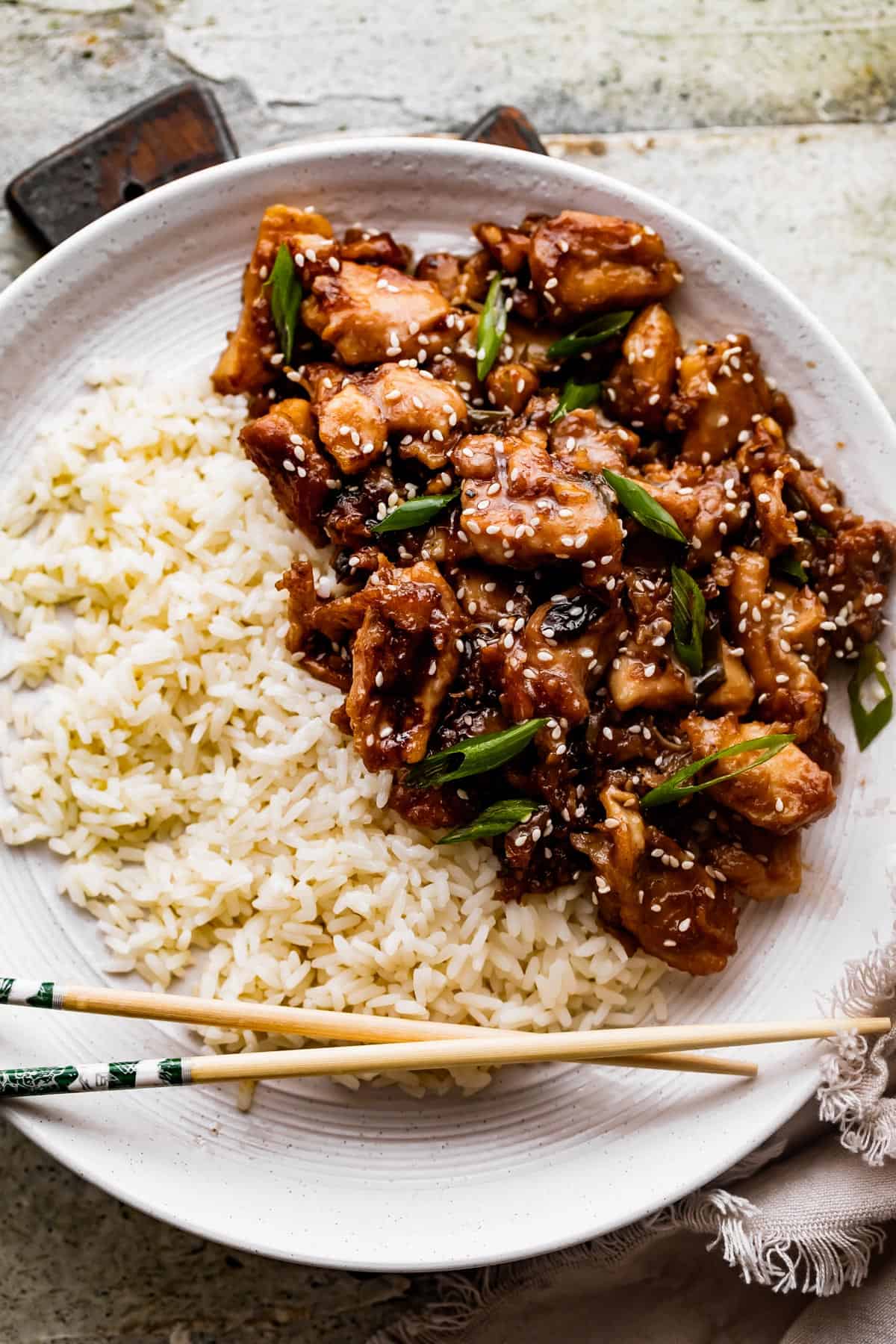 overhead shot of a white dinner plate with bourbon chicken and rice arranged on top of it, and a pair of chopsticks set at the bottom of the plate.