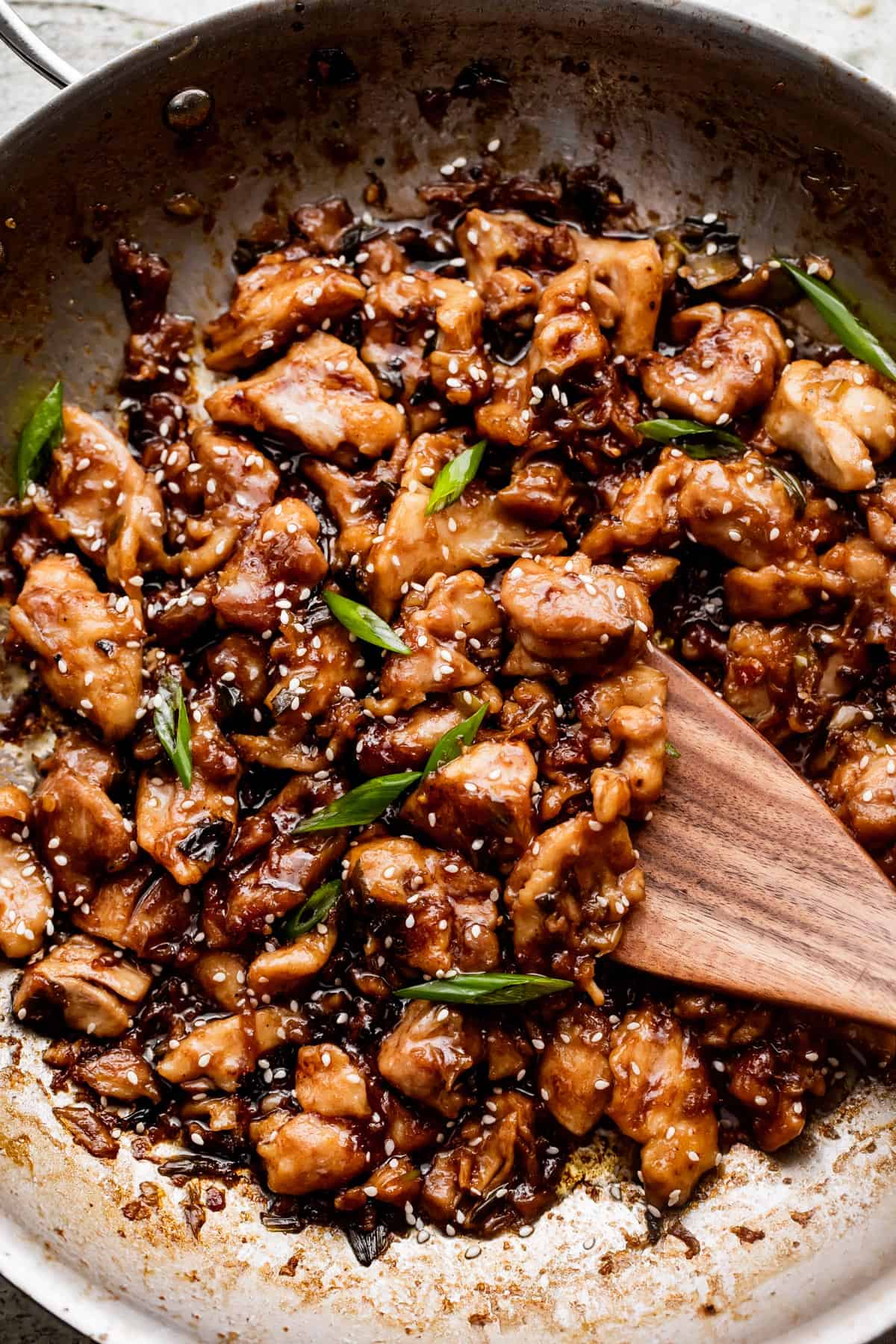 wooden spoon stirring through bourbon chicken while cooking in a stainless steel skillet.