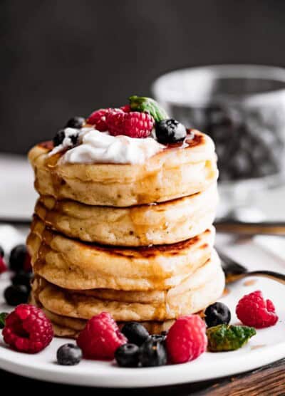 a stack of four drop scones set on a plate and topped with whipped cream, blueberries, raspberries, and a drizzle of pancake syrup.