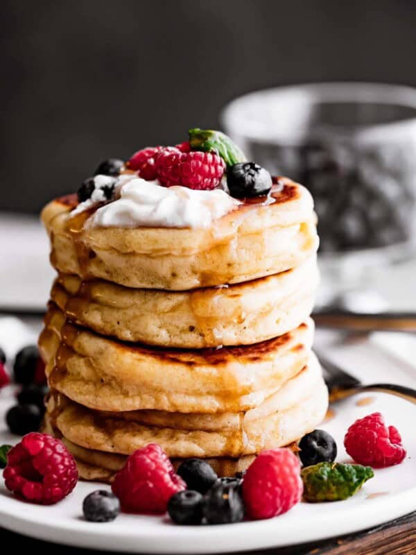 a stack of four drop scones set on a plate and topped with whipped cream, blueberries, raspberries, and a drizzle of pancake syrup.