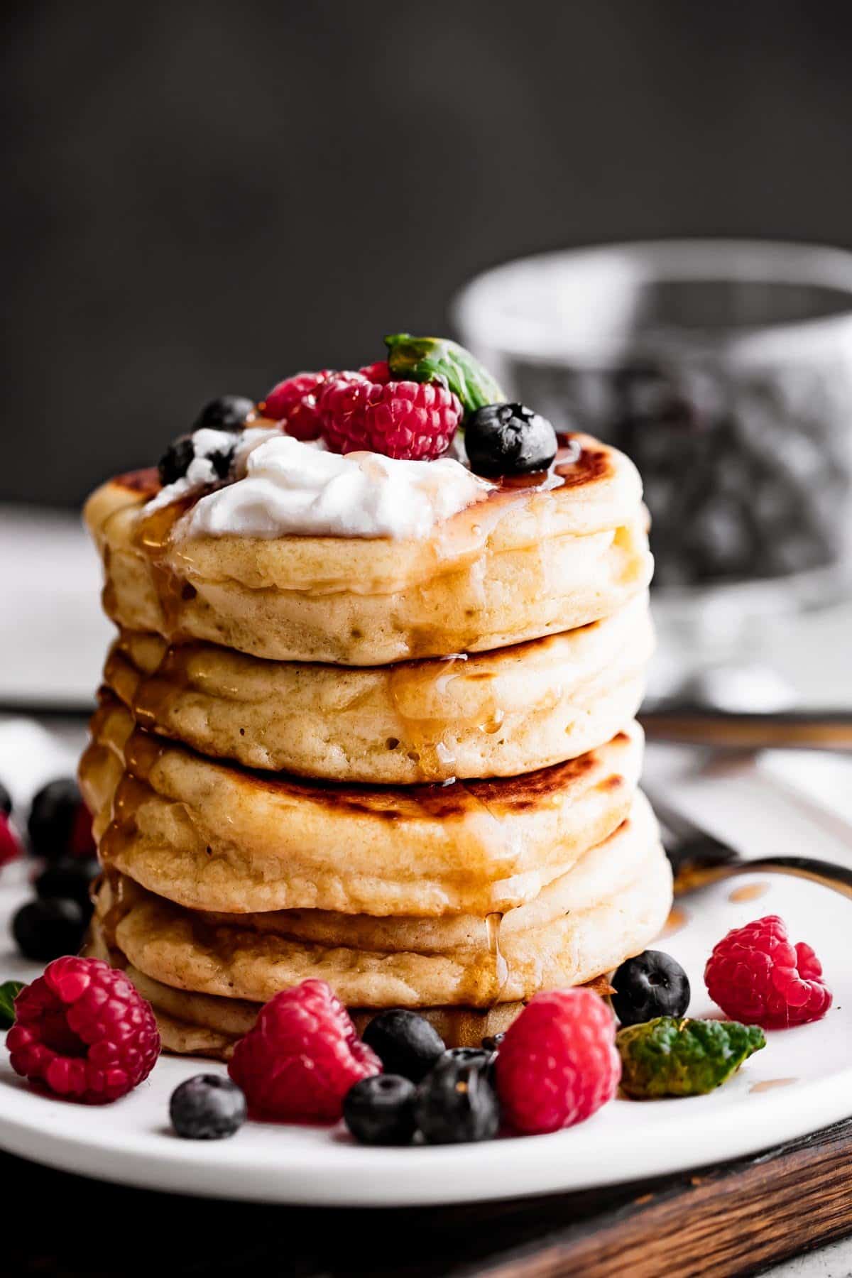 A stack of four drop scones set on a plate and topped with whipped cream, blueberries, raspberries, and a drizzle of pancake syrup.