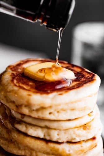pouring pancake syrup over a stack of drop scones topped with a pat of butter.