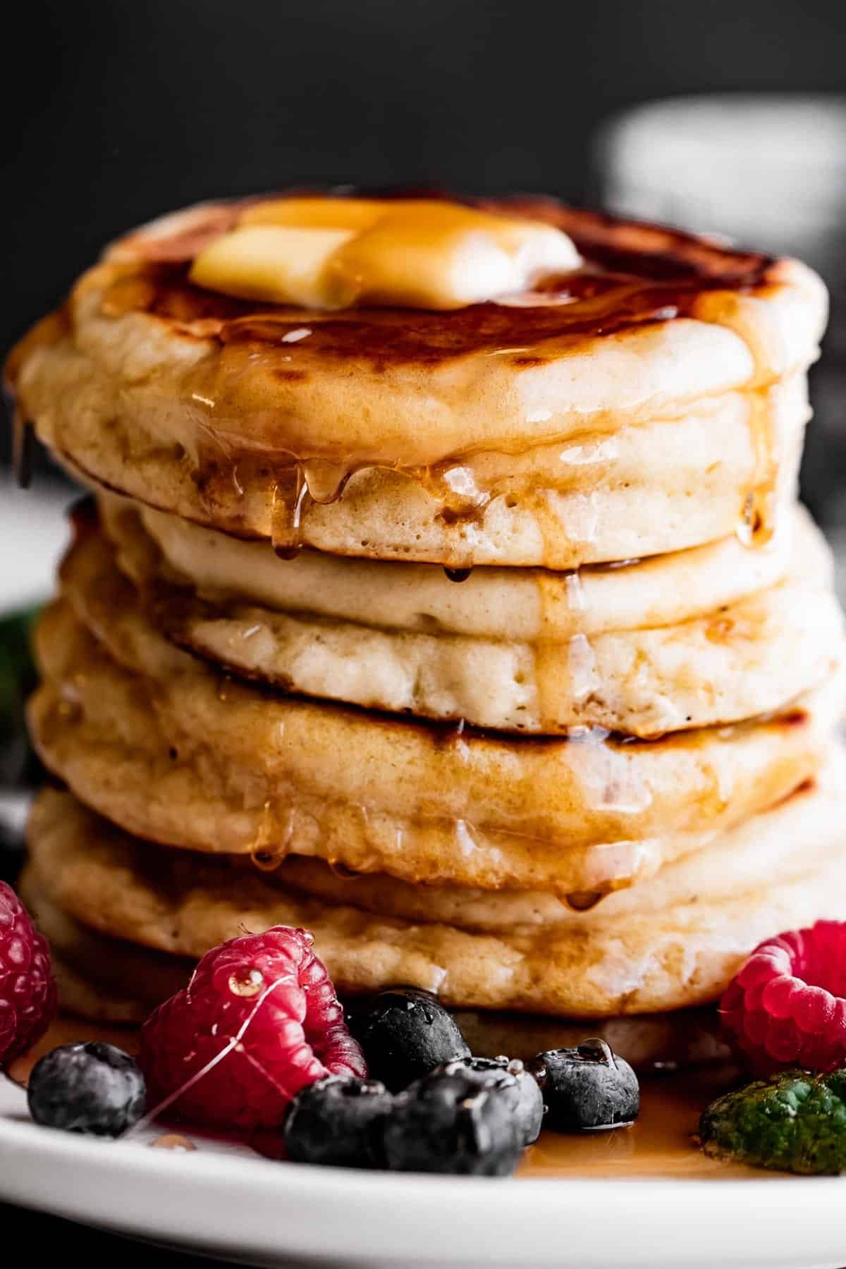 up close shot of a stack of drop scones with a pat of butter on top, drizzling pancake syrup on the sides, and blueberries and raspberries arranged around the scones.