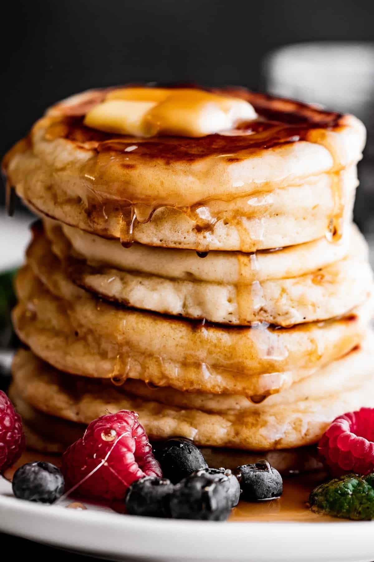 Close-up shot of a stack of drop scones with a pat of butter on top, drizzling pancake syrup on the sides, and blueberries and raspberries arranged around the scones.