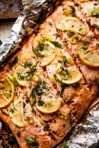 close up shot of a whole cooked salmon fillet set on a piece of foil and topped with lemon slices, herbs, and green onions.