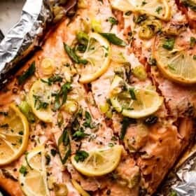 close up shot of a whole cooked salmon fillet set on a piece of foil and topped with lemon slices, herbs, and green onions.