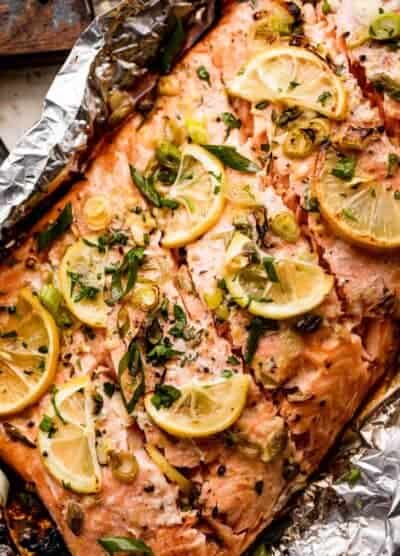 close up shot of a whole cooked salmon fillet set on a piece of foil and topped with lemon slices, herbs, and green onions.