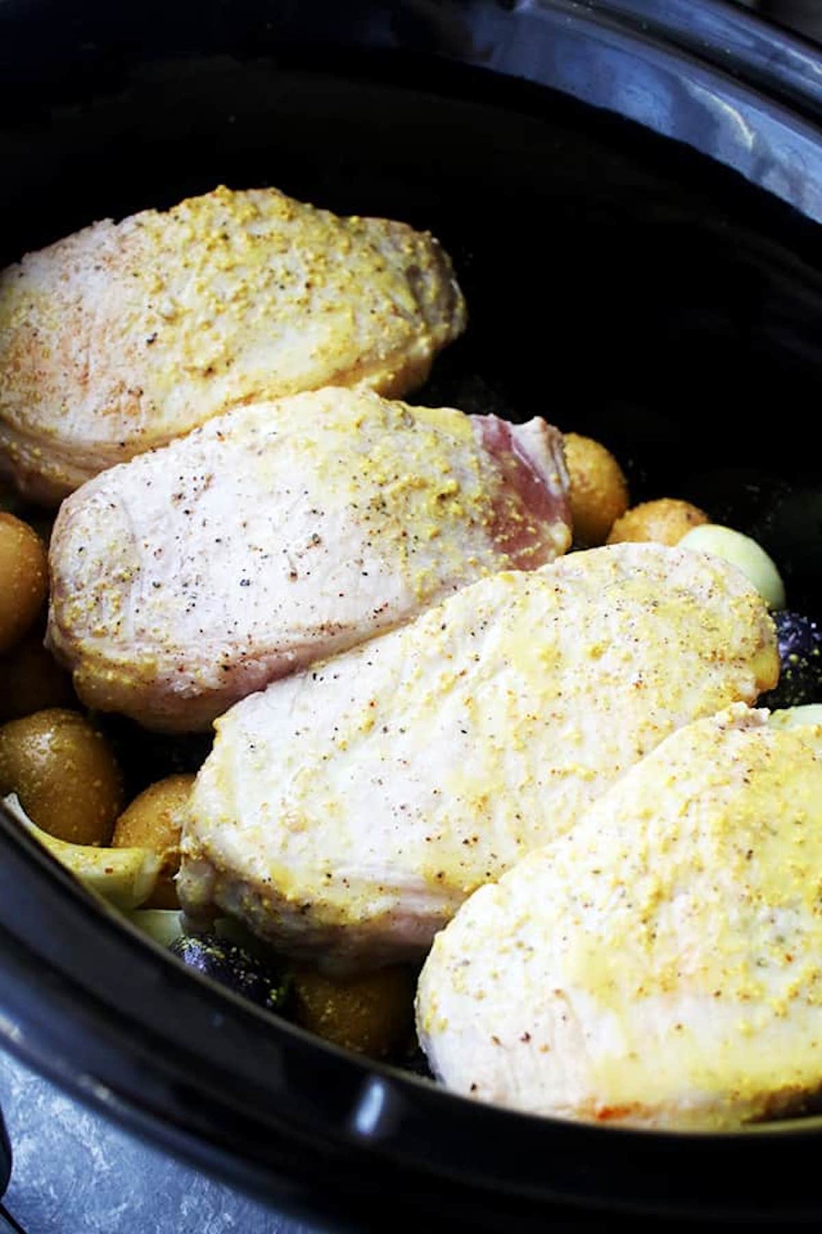 Pork chops in a crock pot with potatoes and onions underneath them and mustard sauce on top of them
