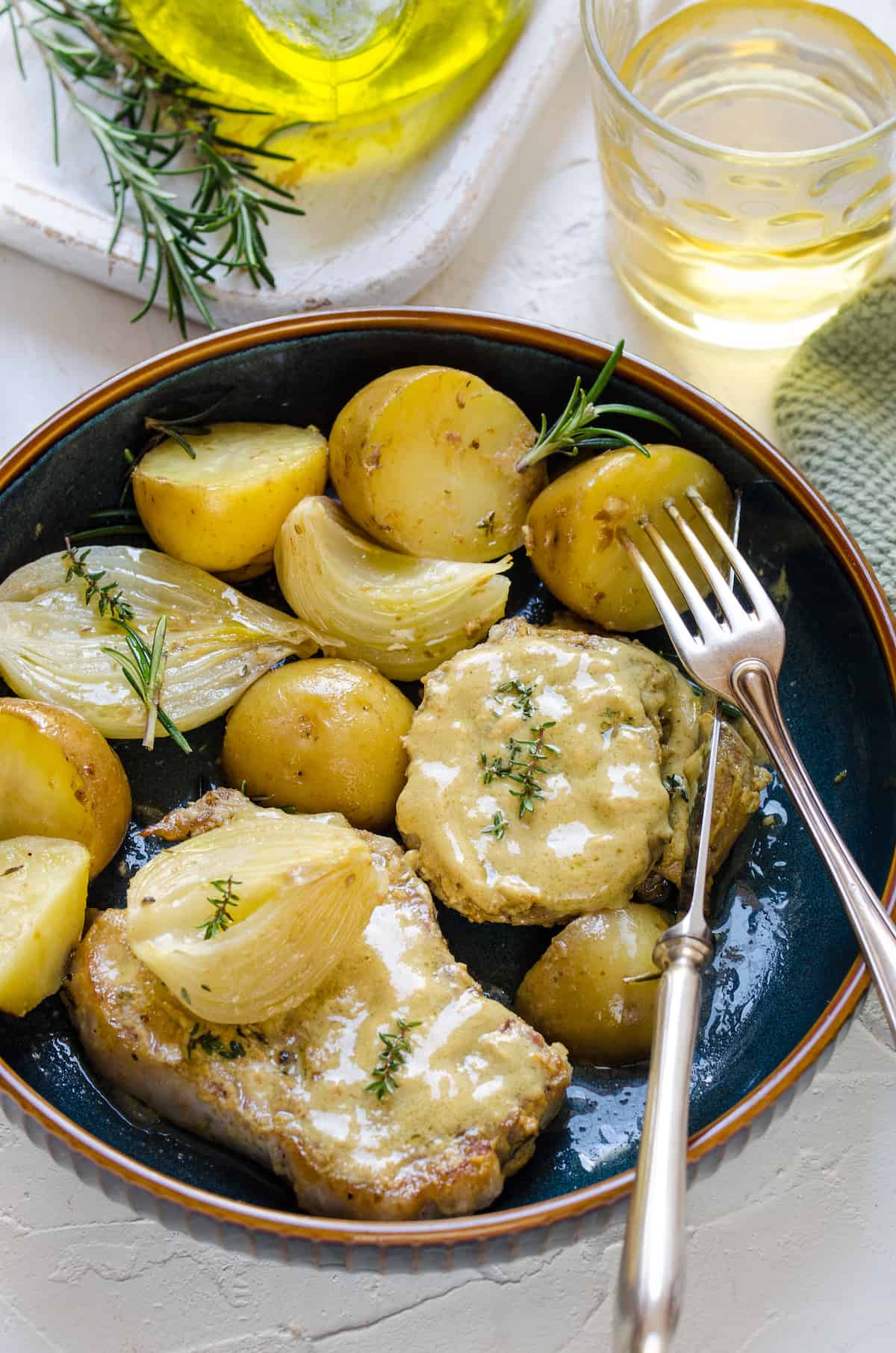 Slow-cooked pork chops, potatoes and onions on a plate beside two sprigs of rosemary and a glass of wine