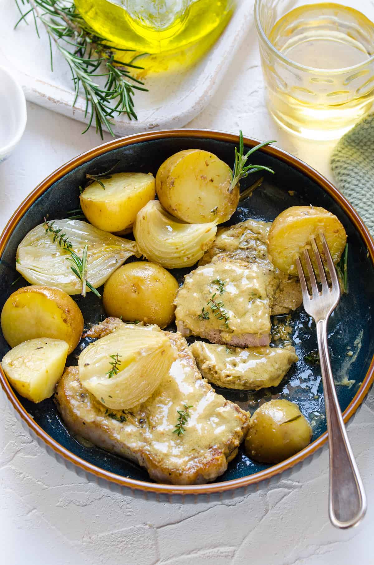 A plate of mustard pork chops on a table beside a glass of white wine and a couple of rosemary sprigs