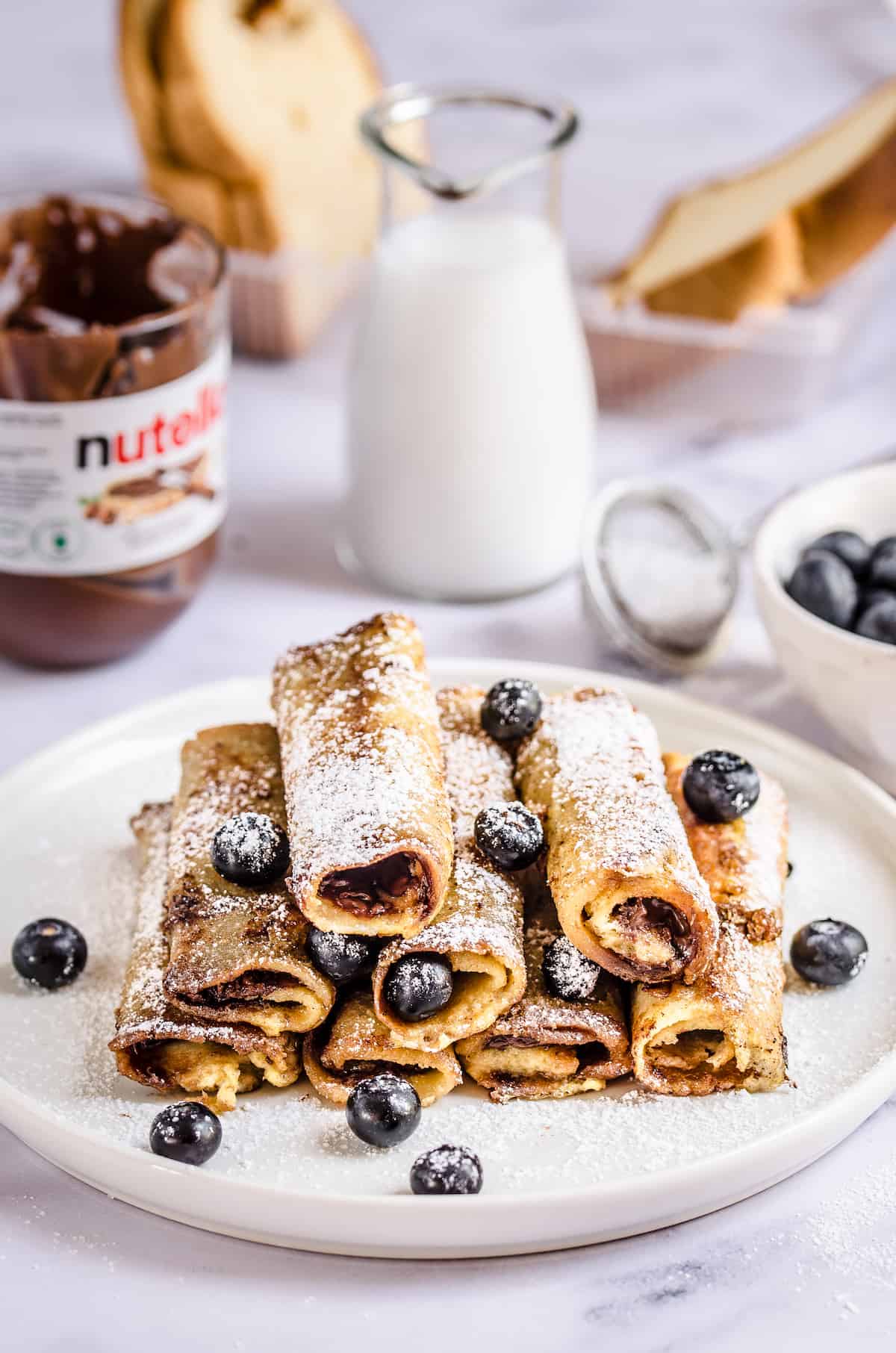 A plate full of Nutella roll-ups with a glass of milk, a bottle of Nutella and a few slices of bread in the background