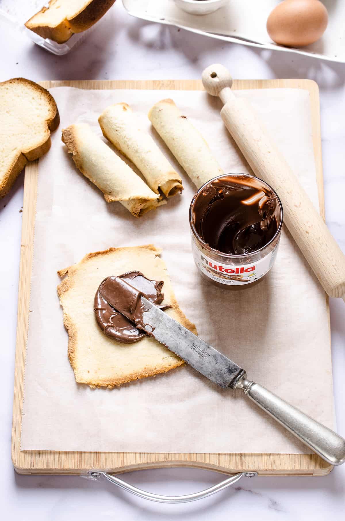 Nutella being spread onto a flattened piece of bread on top of a cutting board