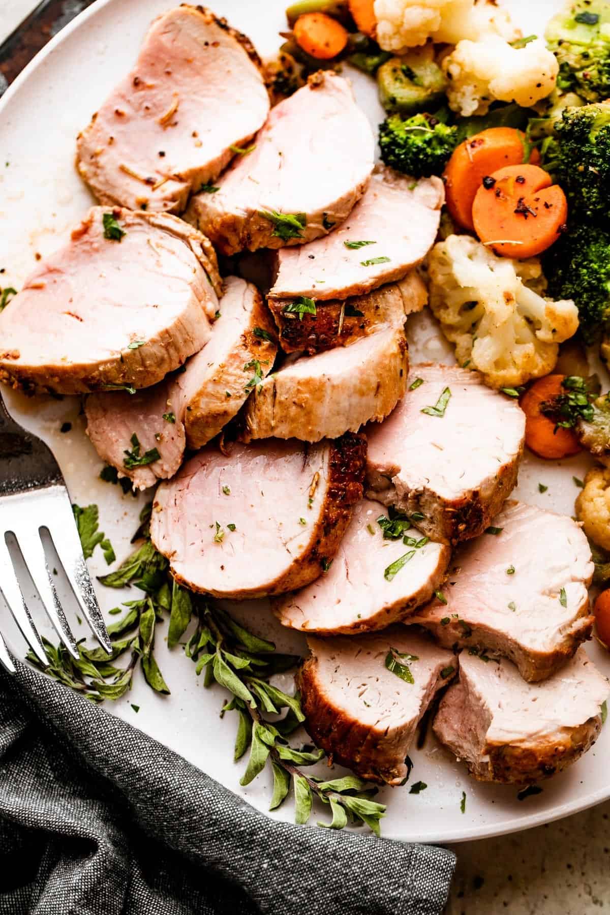 Overhead shot of Air Fryer Pork Tenderloin slices arranged on a white plate with cooked vegetables placed next to it on one side, and a large fork on the other side.