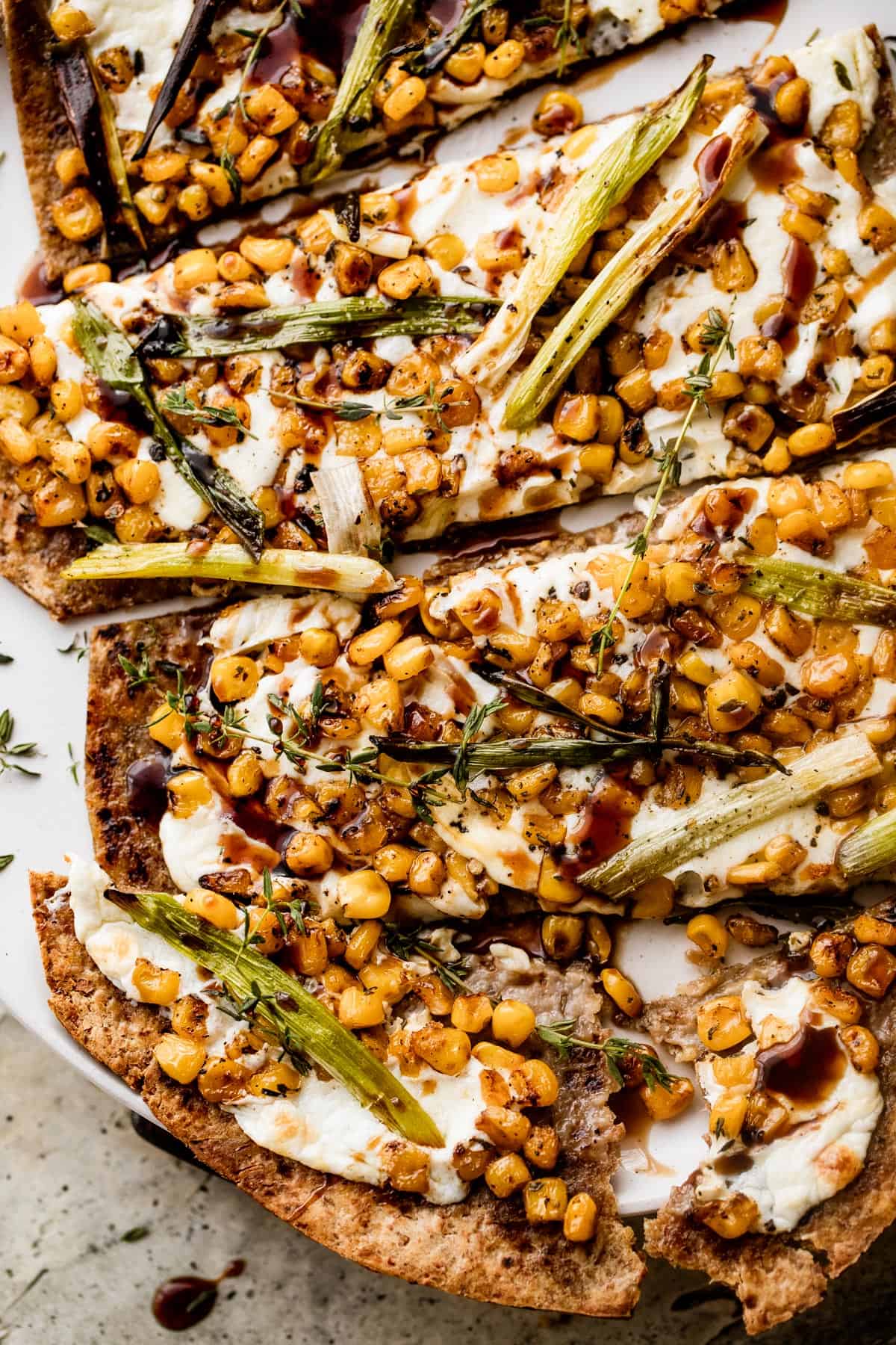 overhead up close shot of a sliced grilled flatbread pizza topped with melted mozzarella, grilled corn kernels, and halved green onions scattered on top, and balsamic