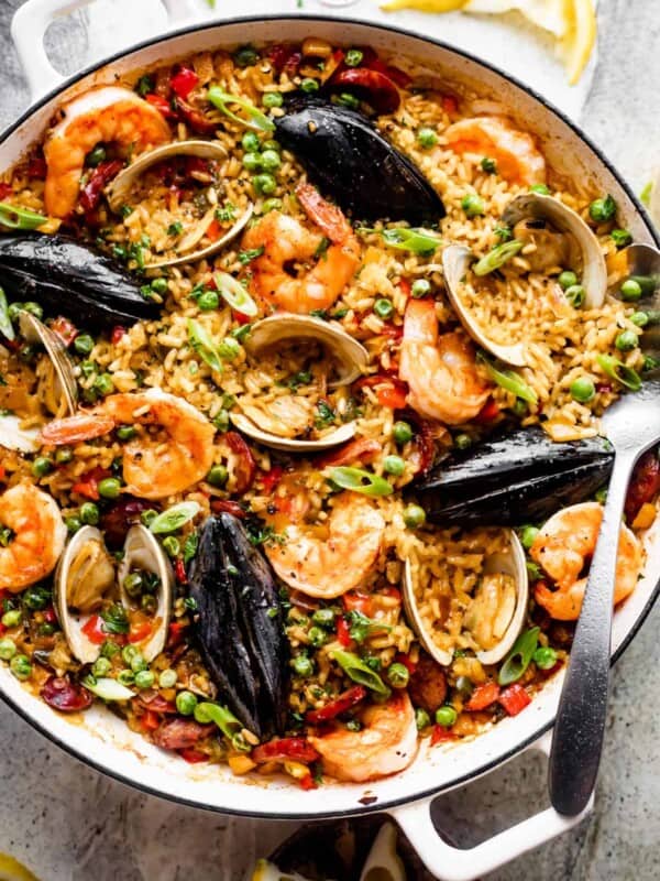 Spanish paella in a white braiser: arborio rice topped with clams, shells, mussels, shrimp, sliced sausages, and peas.