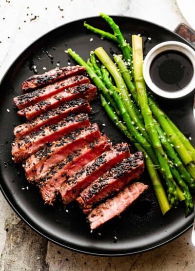 Sliced pan seared sesame crusted tuna steak and served on a black dinner plate with a side of asparagus and soy sauce in a bowl for dipping.