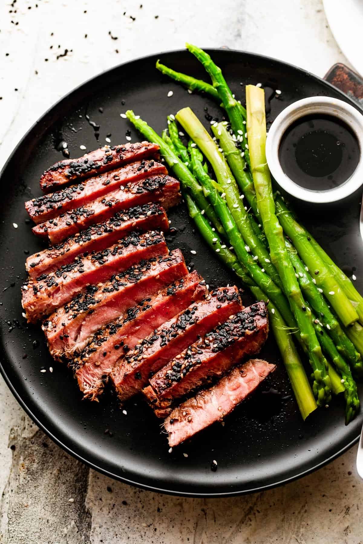 Sliced pan seared sesame crusted tuna steak and served on a black dinner plate with a side of asparagus and soy sauce in a bowl for dipping.