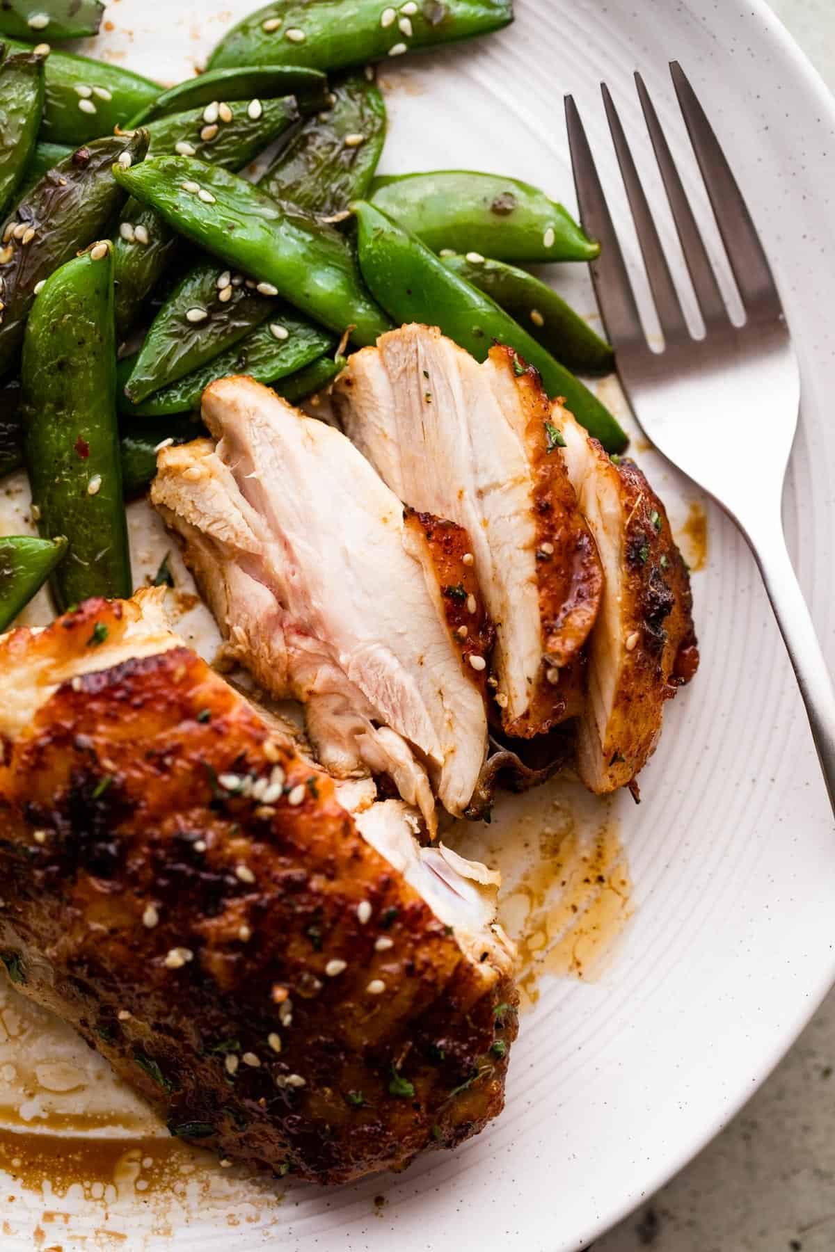 cooked and sliced chicken thigh on a white dinner plate, served alongside sugar snap peas, and a fork placed next to the chicken.