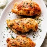 overhead shot of three chicken breasts arranged on a long oblong serving dish, with utensils placed to the right of the plate, and a jar of basil pesto plus a jar of crumbled feta placed to the left of it.